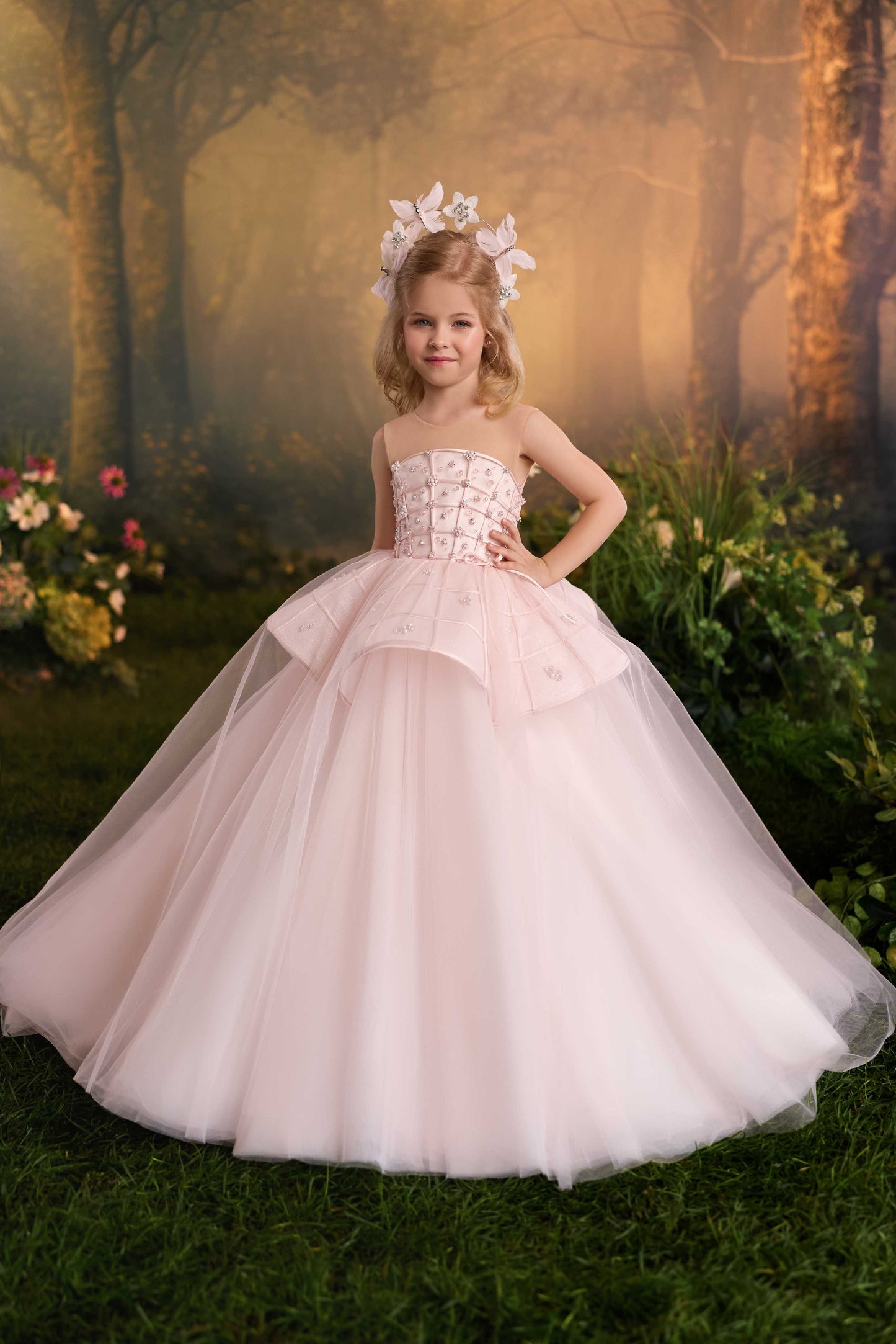 Young girl wearing a soft blush pink princess dress featuring a flower-embellished bodice, tiered peplum overlay, and full tulle skirt.