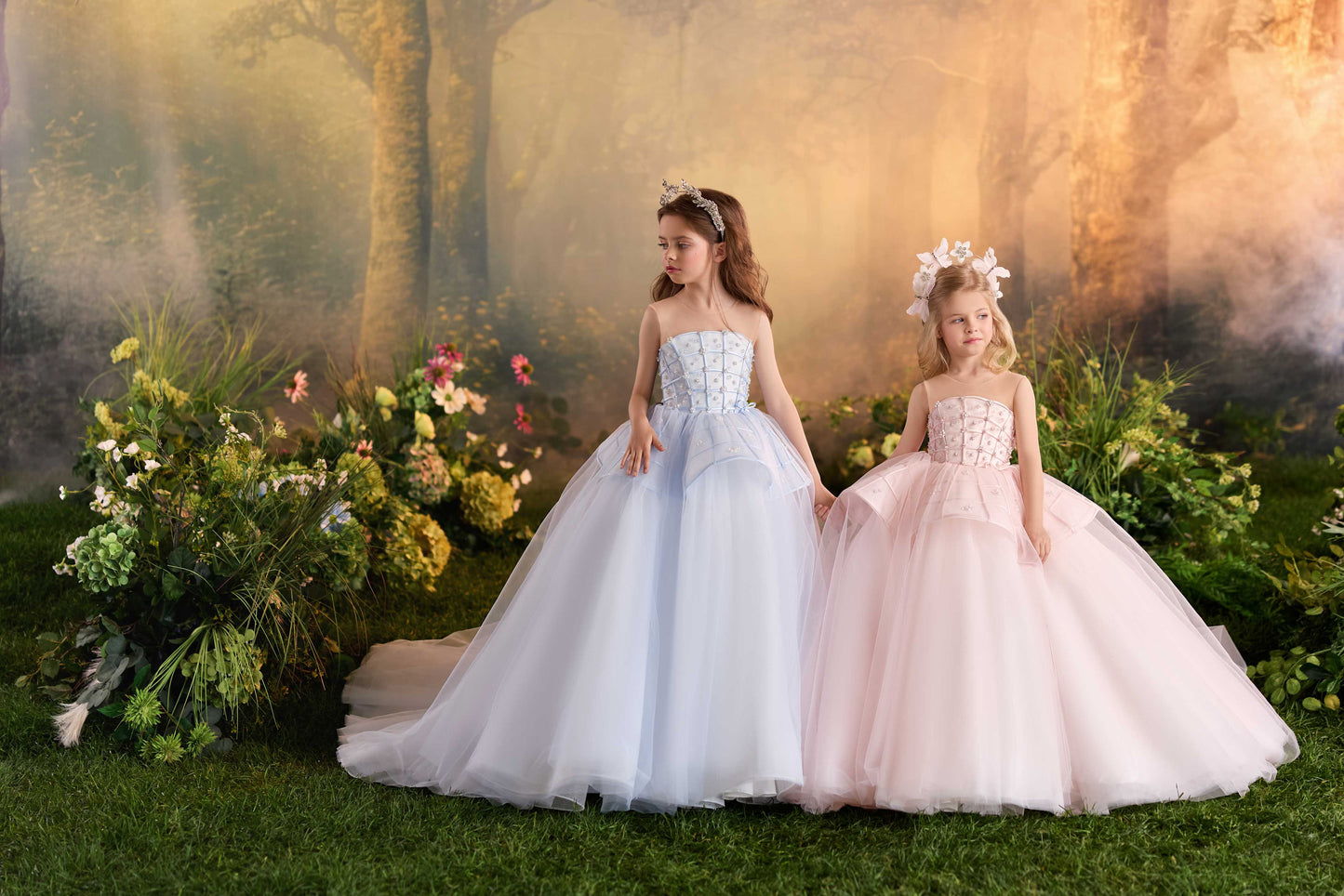 Two young girls standing hand-in-hand modeling luxury strapless-style tulle ball gowns in pastel blue and blush pink against a fairytale forest background